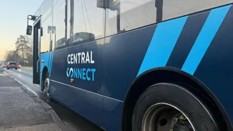 Robyn Wallis/BBC The side of a blue bus parked at a bus stop. Its door is open. The logo of the bus operator Central Connect is printed on the bus in blue and white lettering.