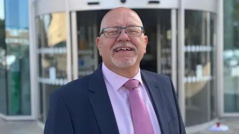 LDRS An older man with a short white goatee beard and glasses smiles at the camera. He wears a navy blue suit with a light pink shirt and darker pink tie. 