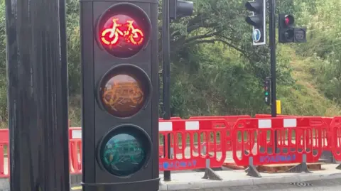 BBC / Samantha Whelan A close up photos of a bicycle traffic light, with the red light illuminated. York Station can be seen reflected in the unlit lights, and there's red plastic barriers by the road behind. There are more traffic lights, and trees and grass in the background.