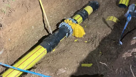 Wales & West Utilities A section of a large yellow and black gas pipeline in a trench. The pipeline includes a joint which appears to be part of a connection or repair. The surrounding area is soil.