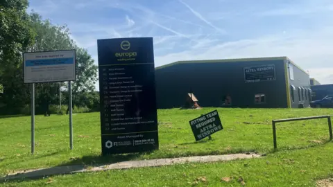 Photograph of the front of the Europa Trading Estate in the Stoneclough area of Bolton. The image shows a sign, with corrugated iron sheds in the background.