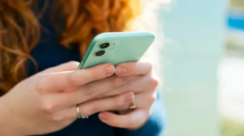 A girl with red hair holds a mobile phone contained in green case. The girl is wearing a blue top.