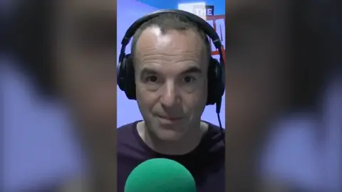 A man wearing headphones looks at the camera, sat in front of a green mic.