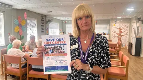 Debs is holding a 'save our day centre' post and members are behind her sitting down. 