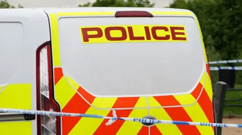 The back of a police van, which is parked near a park, with police tape in front of it. 