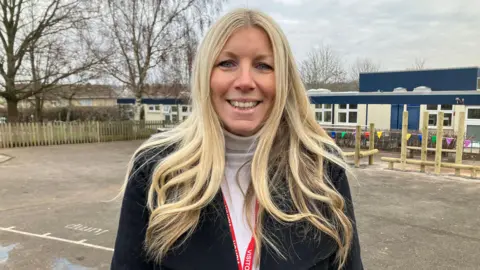 Charlotte smiles into the camera. She has long blonde hair and is wearing a black coat with a white polar neck jumper. She is standing in the school playground with the new equipment behind her. 