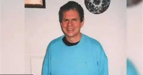 A man, Stephen Ridgeway, is smiling at the camera and wearing a blue jumper. He has short dark grey hair.