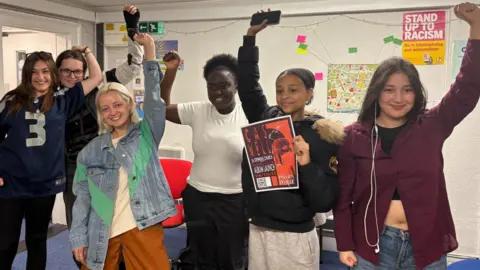 Young women are celebrating the news of their funding with their arms in the air. There are six of them, one is holding a poster, another has headphones hanging out of her ears. They are in a room with a white wall covered in posters. 