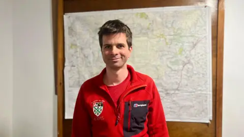 Plymouth College leader Olly Rees smiling at the camera and stood in front of a map. He is wearing a red fleece with the school badge on the left. 