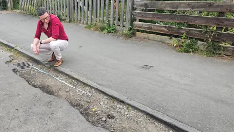 LDRS A man in a red shirt and white jeans crouched down measuring a pothole with a measuring tape. He is on the pavement and his measuring tape is laid out on top of a large pothole in the road. 
