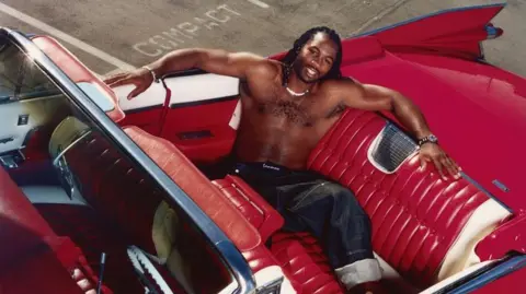 Steve Read A portrait photograph of Lennox Lewis sitting in the back of a red convertible car with the roof down. The vehicle is parked on a street and the boxer is photographed from above.
