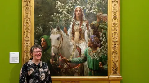 Bradford Museums and Galleries A person with short hair and a colourful shirt stands next to a painting in a gallery. The painting shows a woman in white on a white horse surrounded by flowers she is being attended by men in green