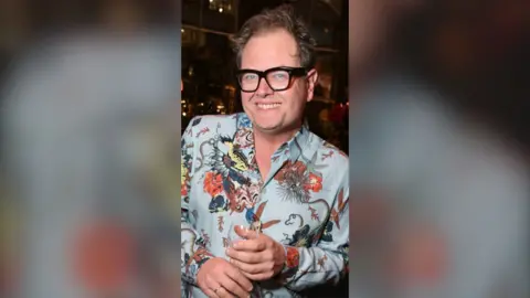 Alan Carr smiles at the camera. He is wearing a pale blue shirt with an elaborate floral pattern on it.