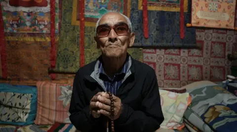 SANDEEP YADAV/BBC Dawa Sangbo with sunglasses sitting and wearing a black jacket over a blue scheme shirt and prayer beads. The wall behind it is covered with colored carpet.