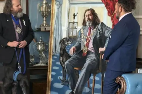 The Mayor of Bournemouth Two men in dark blue suits one on the left with a greyhound dog on a lead stand either side of a large portrait.