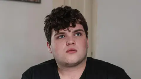 Dylan Cullen poses for picture while sat on a sofa. He wears a black Vans shirt and white walls are behind him.