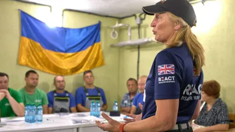 Lewis Inman A woman with long blond hair and a dark baseball cap talks to a room of people sitting behind a table. There is a large Ukranian flag on the wall behind them and several bottles of water on the table. The woman has a Union Jack flag on her sleeve.