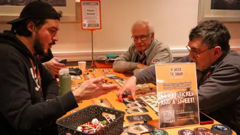 Paul Harris A group of men of various ages are sat at a table covered in cards and other board game paraphernalia