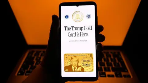Getty Images The Trump Gold Card website is displayed on a mobile phone screen posed in front of a laptop with an orange background. The Gold Card webpage says "The Trump Gold Card is Here" with the subtitle, "Unlock life in America" and an image of the card, with Trump's image