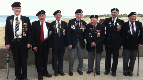 Olivia Richwald/BBC Seven older men wearing suits with many military medals and badges on them are lined up with a beach in the background. Some of them are holding walking sticks.