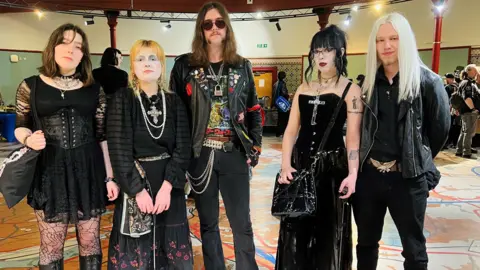 Three women and two men stood in row looking straight ahead. They are all wearing black clothing. The men have long hair while two of the women are wearing heavy make-up. They are stood in a large room with people in the background. 