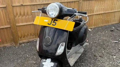 Simon Drieu A black scooter is parked on gravel next to a wooden fence. It has the registration plate attached to its handlebars - it reads J5.