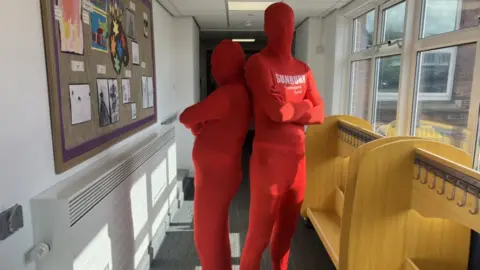 Lara King / BBC Two men standing back to back dressed in head to toe bright red Morphsuits. Their whole bodies are covered in the red suits. On the chest of the suits it says Sunburn. They are standing in a school corridor with waist high racks with coat hooks on the right and a notice board on the left.
