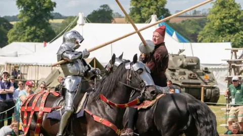 Chalke History Festival Men on horseback, one in full armour. They are re-enactors, the horses are wearing traditional tack for a demonstration of fighting with lances or jousting. A fence surrounds, which spectators are watching from with the roofs of numerous marquees in the background