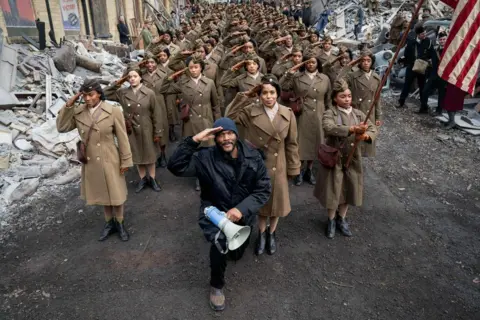 Netflix Tyler Perry on set in a woollen hat and black coat, carrying a megaphone, in front of a group of women in period military uniform, one carrying a US flag. Everyone is saluting.