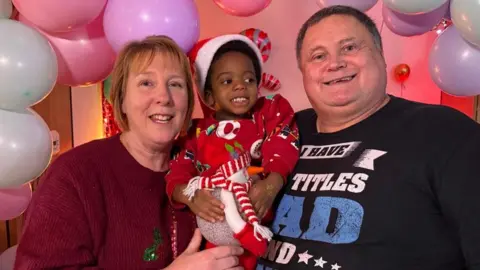 A woman with blonde hair is wearing a red top and holding a child dressed in a Santa outfit. On the right is a man with a black top smiling.