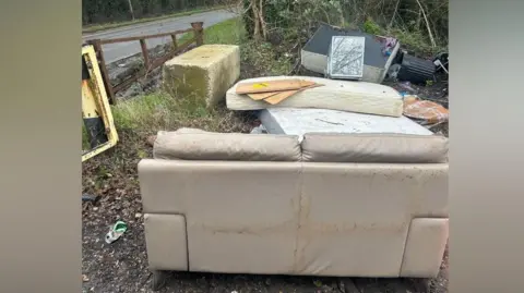COLIN RAYNER Fly-tipping on land next to a road. A sofa, mattresses and other rubbish are near a concrete block and a small gate.
