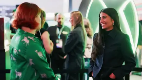 A woman with long dark hair dressed in all black is smiling while chatting to another woman with short red hair dressed in green.