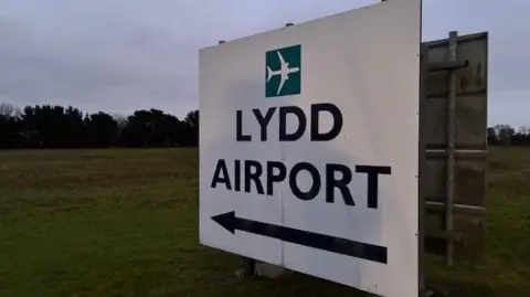 Simon Jones/BBC A white sign in a grass field. The sign reads Lydd Airport, has a large arrow to the left and bears a small airplane icon.