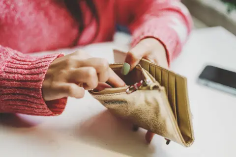 Getty Images A close-up of a woman's hands. She wears a pink sweater and rummages through a gold purse for money. 