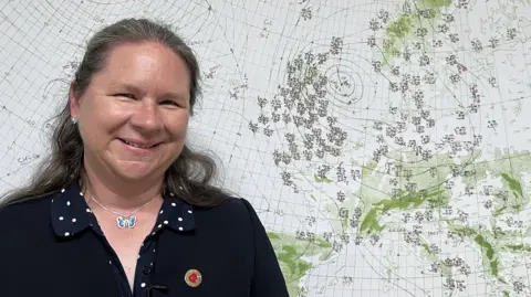 Dr Catherine Ross has dark grey long hair, and is wearing a dark jacket and a navy and white polka dot dress. She is smiling while standing in front of a hand-drawn weather chart from D-Day in 1944.
