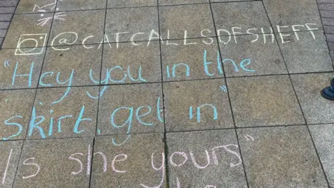 Cat Call Sheffield A chalked pavement, the work of Cat Calls Of Sheffield, part of the 'chalk back' campaign where cat calls are written in large cursive handwriting in chalk, in the area of the city they took place. The words "Hey you in the skirt, get in" are written on a pavement in blue chalk, underneath the Instagram hashtag @catcallsofsheffield written in pale yellow chalk, and in the top left hand corner of the text is a small cat image of eyes, ears and whiskers drawn in pink chalk.