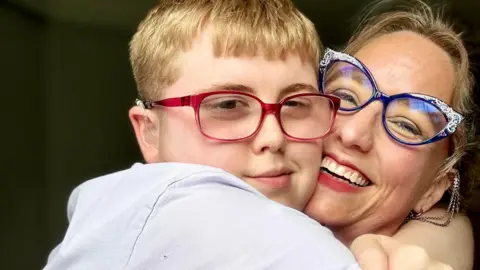 Family picture Adam hugs his mother tightly and she smiles into camera. He wears a white top and red-rimmed glasses and has short blonde hair. She smiles showing her teeth and has blonde hair, pulled behind her head, and blue, horn-rimmed glasses with sparkly edges.