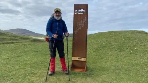 Charles Bain Smith stands beside the Hebridean Way sign in outdor walking gear and holding walking poles, with the Scottish coastline and mountains in the background.