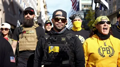 Getty Images Tarrio successful  sunglasses and a vest containing 2  yellowish  cans, flanked by others wearing Proud Boys gear