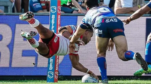 St Helens wing Lewis Murphy scoring a try for his side by diving for the line with his left hand on top of the ball which is grounded on the line while two Wakefield defenders try to stop him