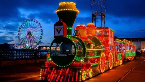 Illuminated heritage tram on Blackpool promenade at night with Central Pier in the background and the Big Wheel lit up.