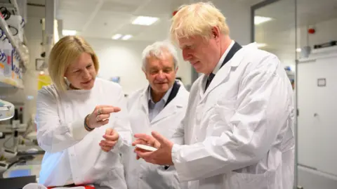PA Three people in white lab coats stand in a bright laboratory. The woman, Lucy Collinson, on the left points at something in her hand. The blond man, Boris Johnson, on the right holds a small object. Sir Paul stands between them, all focused on the discussion.