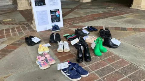 A group of children's shoes in pairs on the ground with labels attach to them.