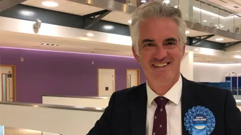 Jon Wright/BBC James Cartlidge wears a suit with a burgundy tie. He is smiling and has a Conservative badge on his left lapel