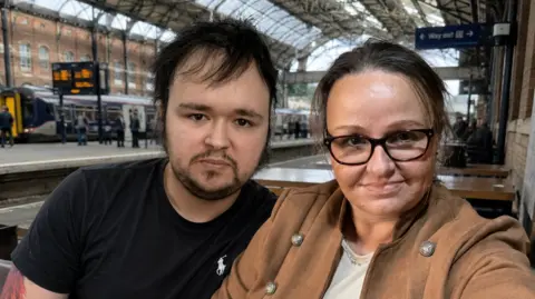 PA Real Life A young man with dark hair and a black t-shirt sits next to an older woman with brown hair and a brown coat. They both look at the camera and sit at a table in a railway station.