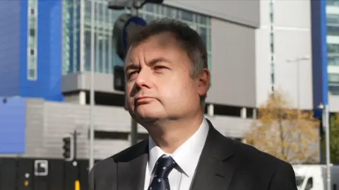 A man in a dark grey suit with a navy tie stands in front of a blue and grey clad building