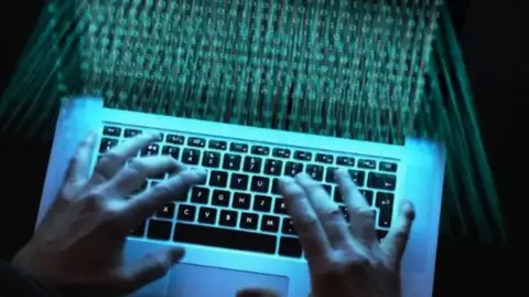 Image of a someone typing on a computer in the dark. Blue light from the screen shines on the person's hands and keys. The screen is blurred but numbers can be seen on the screen. 