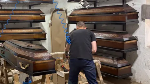 The back of a man using power tools to make coffins. There are around half a dozen identical coffins surrounding him stacked on the wall. 