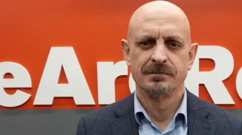 Dawid Wojtowicz/BBC Dave Perry. A man who has a moustache and is looking into the camera. He is dressed in a blue shirt and suit jacket. He is sat down and behind him is a grey and orange backdrop. On the orange backdrop it says WeAreRebels