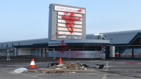 CPS A pile or rubble in front of a grey security building, which is splattered in red paint, and beyond it a large tall sign with company names obscured by a large amount of red paint.